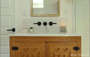 A bathroom vanity from an award-winning remodel featuring an ornate wood cabinet, white countertop, mounted sink, black fixtures, a small plant, a candle, and a mirror above.