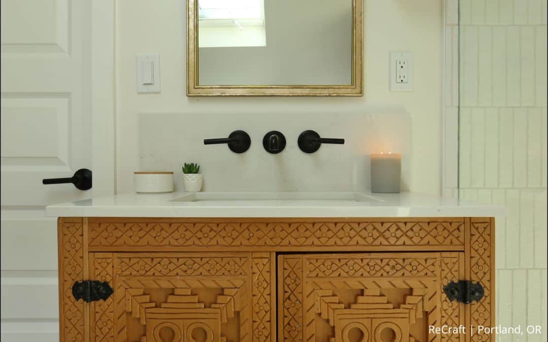 A bathroom vanity from an award-winning remodel featuring an ornate wood cabinet, white countertop, mounted sink, black fixtures, a small plant, a candle, and a mirror above.