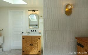Bathroom with white walls, a wooden vanity with decorative carvings, a wall-mounted faucet, and a modern sconce above textured tile. Created by Remodeler of the Year NARI winner. 