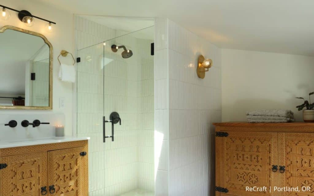 A modern bathroom with a glass shower, wood vanity with ornate carvings, stacked towels, and wall-mounted light fixtures highlights an impressive before and after transformation.