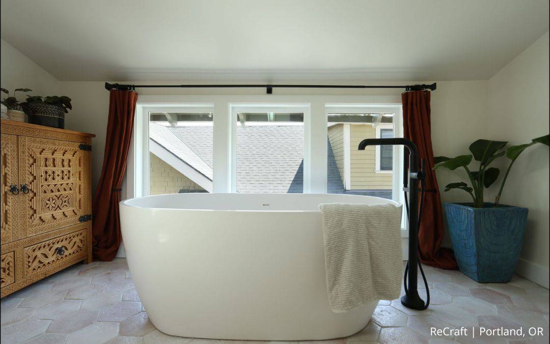 A modern bathroom featuring a freestanding white bathtub, black faucet, towel, carved wood cabinet, large windows, and a potted plant in a blue pot—part of an award-winning remodel by the remodeler of the year.