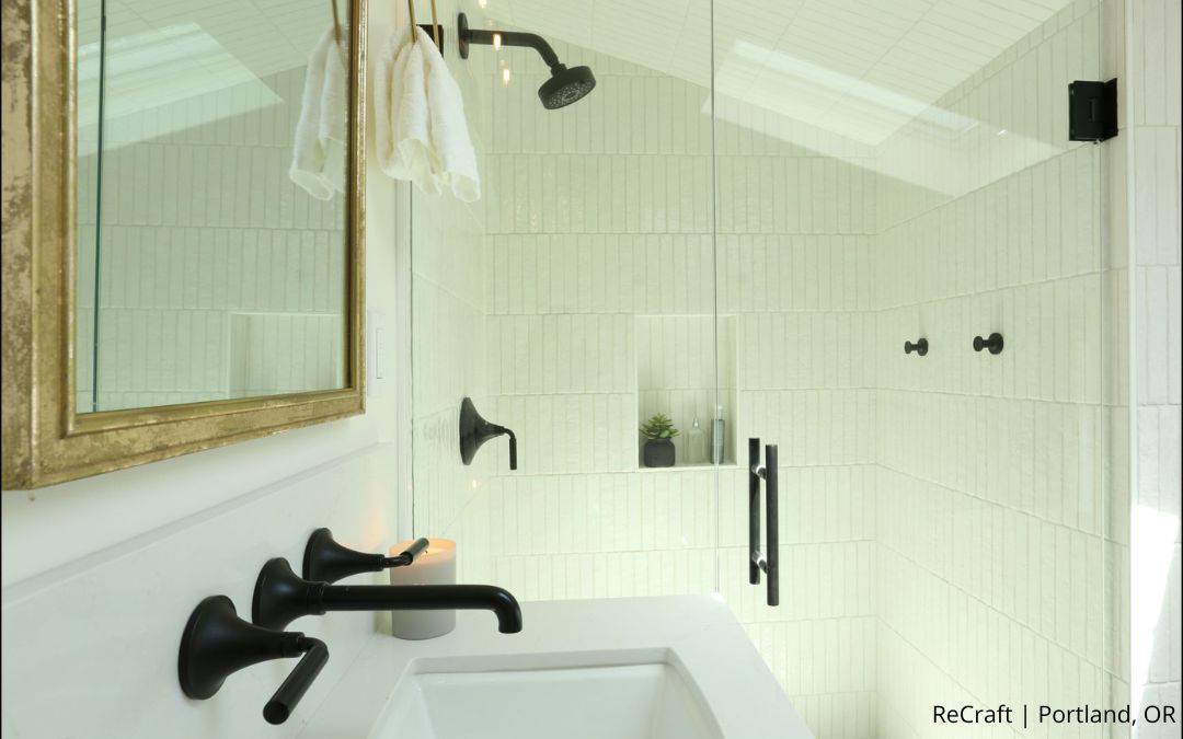 Modern bathroom with white tile, black fixtures, a glass shower enclosure, and a wall-mounted mirror; an award-winning addition by a NARI Remodeler of the Year. Two white towels hang near the shower.