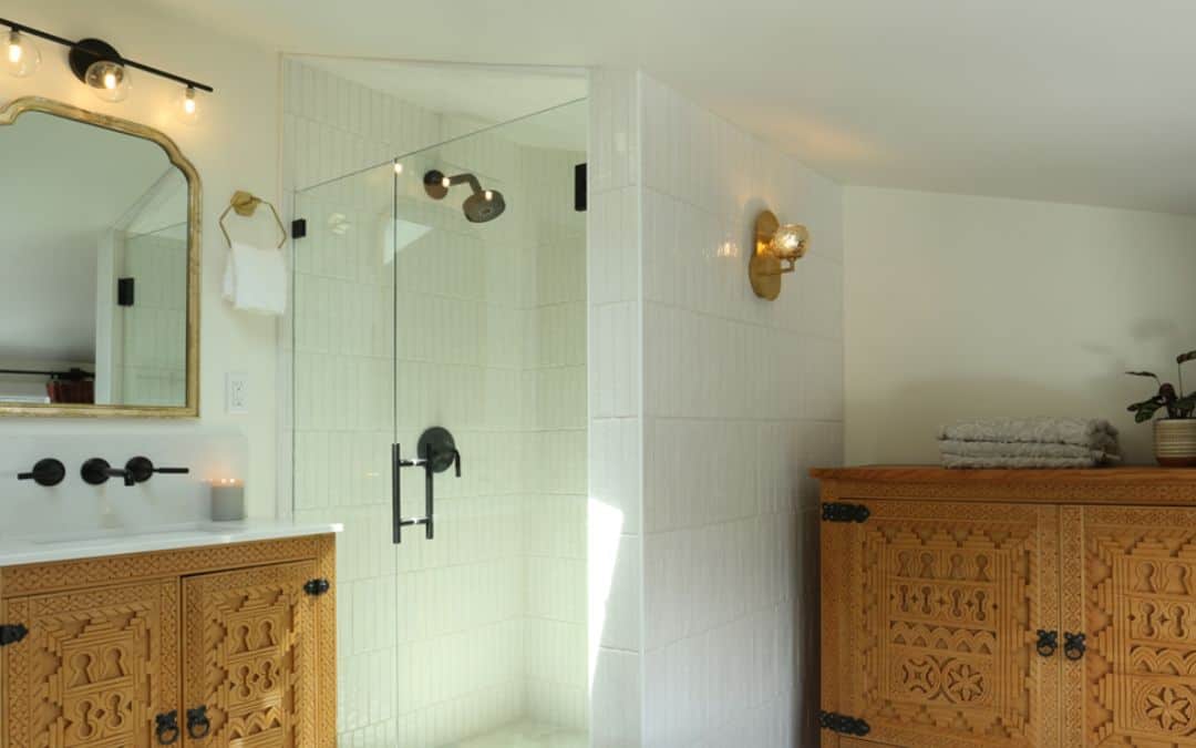 This award-winning addition features a bathroom with a glass shower enclosure, wooden cabinets with carved designs, a mirror, wall lights, and neatly folded towels.