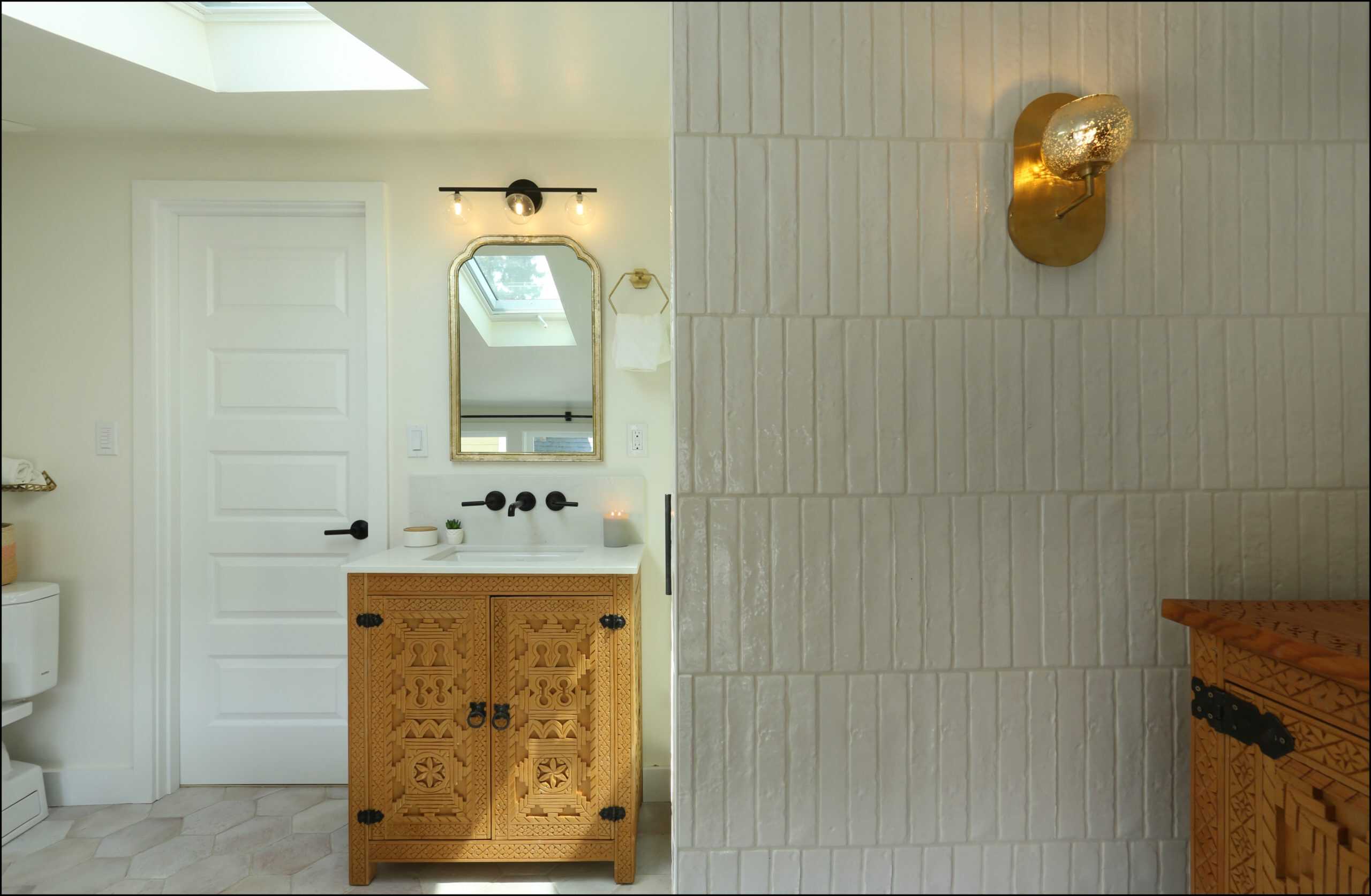 A bathroom with a carved wooden vanity, white sink, rectangular mirror, white tiled wall, and brass wall sconce creates an elegant atmosphere in this NARI award-winning addition.