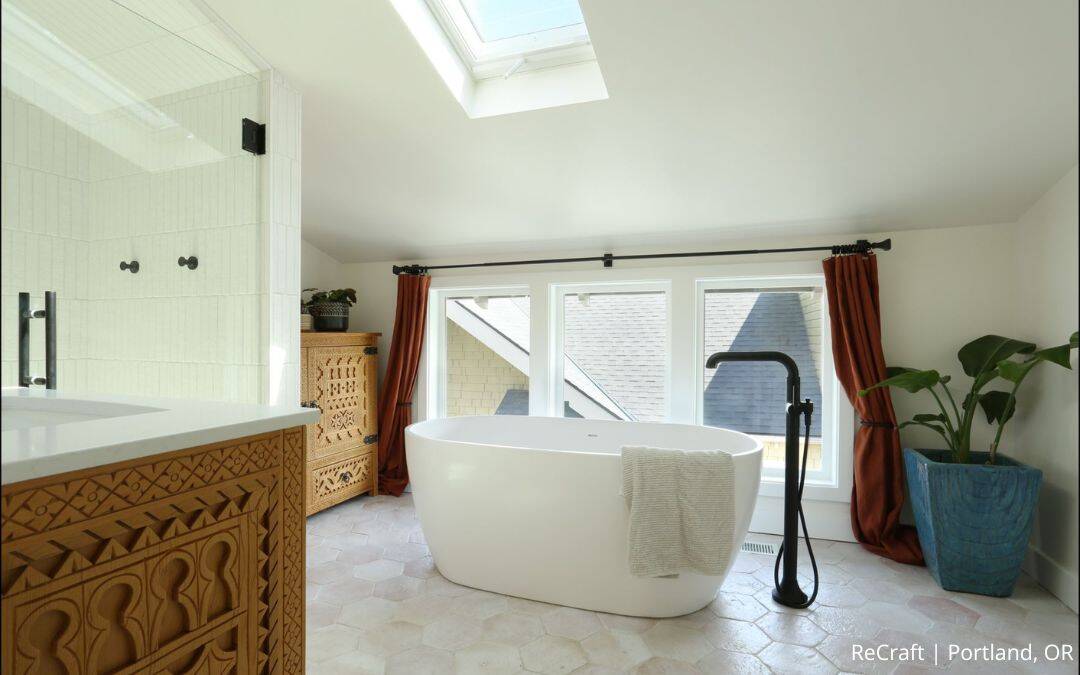 A modern bathroom with a freestanding white tub, black faucet, skylight, large windows with red curtains, wooden cabinet, and potted plant—an award-winning remodel that beautifully balances style and function.
