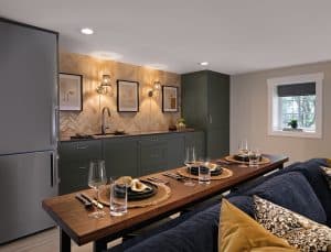 Modern kitchen with dark green cabinets, a wooden countertop, and a dining table set for four. Herringbone tile backsplash, wall art, and soft lighting complete the space.