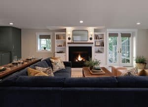 A modern living room with a dark blue sectional sofa, a fireplace, built-in shelves, and glass doors leading outside. The room is decorated with plants and neutral-toned accents.