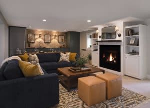 Modern living room with a dark sectional sofa, wooden coffee table, two tan ottomans, a fireplace, built-in shelves, and an open kitchen in the background.