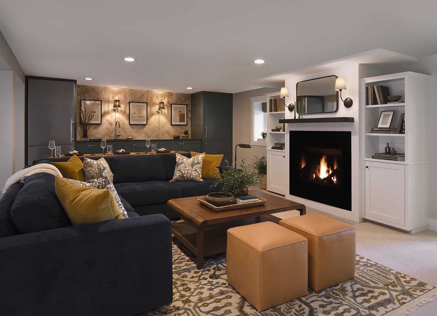 Cozy basement living space featuring a dark blue sofa, decorative pillows, a wooden coffee table, and two tan ottomans, with a fireplace and stylish shelving in the background, showcasing ReCraft Home Remodeling's basement transformation expertise.