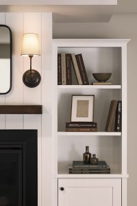 White built-in shelves beside a fireplace display neatly arranged books, a framed picture, decorative bottles, and a bowl under a wall-mounted sconce.