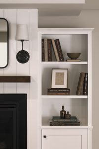 White built-in bookshelf with assorted books, a small bowl, framed art, and decorative bottles next to a fireplace and wall sconce.