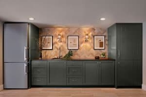 Modern kitchen with dark green cabinets, stainless steel refrigerator, wall art, and decorative lighting on a herringbone backsplash wall.