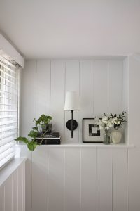 A small ledge holds a potted plant, stacked books, a wall sconce, a framed geometric print, and a vase of white flowers beneath a window with white blinds.