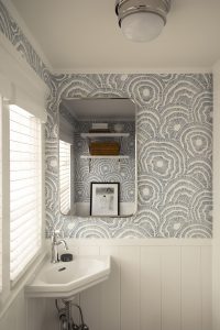 A small bathroom features patterned wallpaper, a rectangular mirror above a corner sink, white paneling below, and a window with white blinds. A shelf and framed picture are reflected in the mirror.