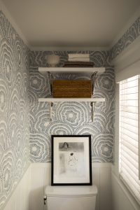 A small bathroom with blue patterned wallpaper, white wainscoting, a window with blinds, shelves holding decor items, and a framed picture resting on the toilet tank.