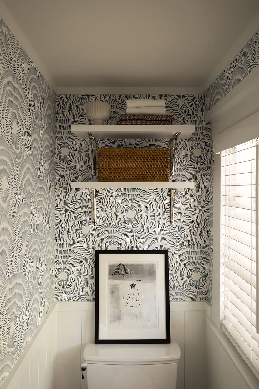 Bathroom corner with decorative wallpaper, white shelves holding a basket and towels, and a framed artwork above a toilet, showcasing ReCraft Home Remodeling's basement design aesthetics.