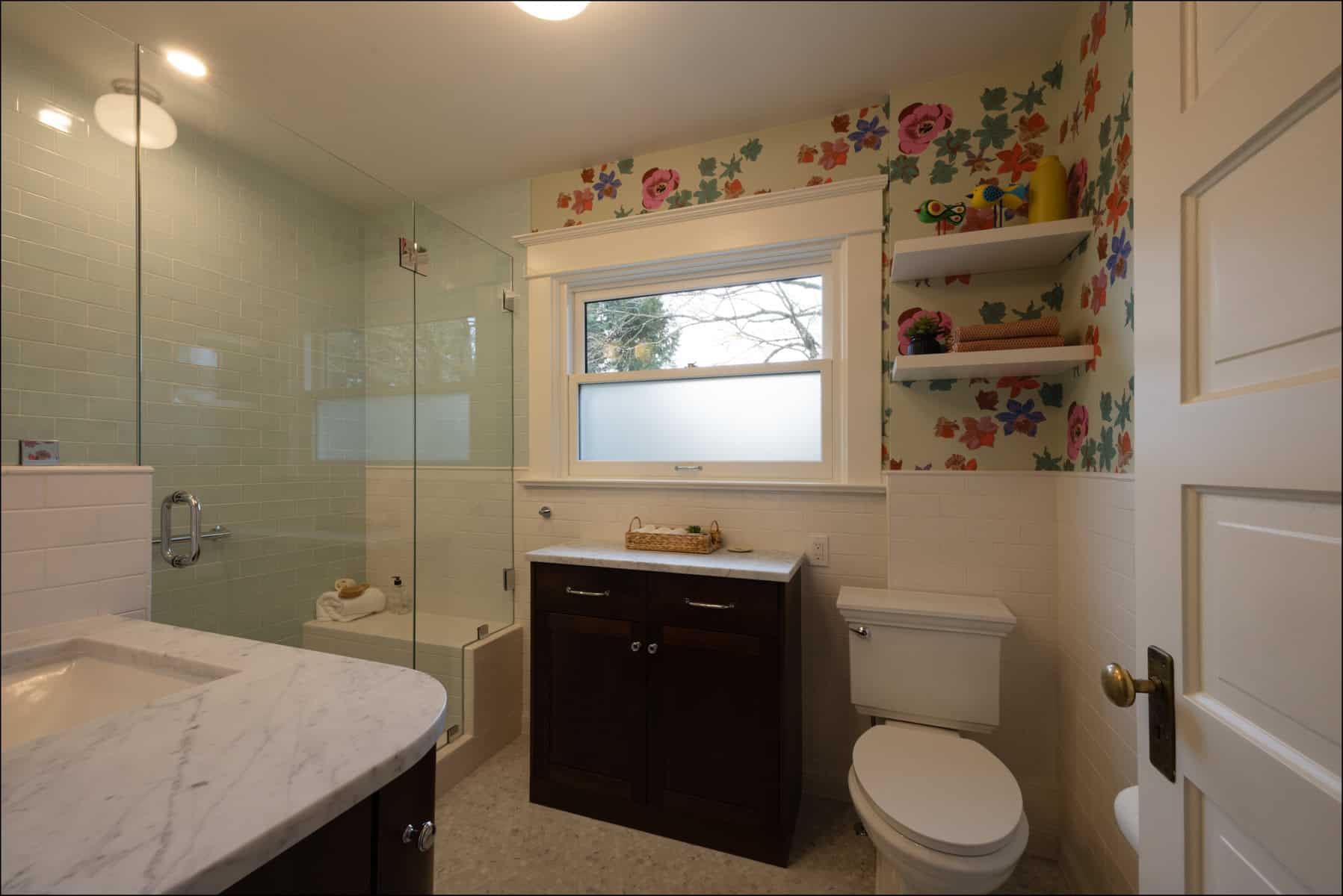 A bathroom with floral wallpaper, glass-enclosed shower, white toilet, dark wood vanity, and shelves with colorful decor under a window.