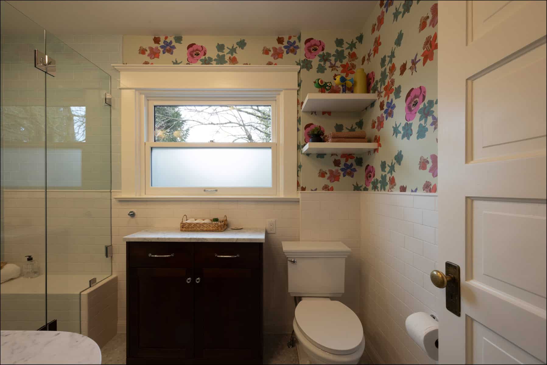 A bathroom with floral wallpaper, a white toilet, a dark wood vanity with a sink, a frosted window, wall shelves, and a glass shower enclosure.