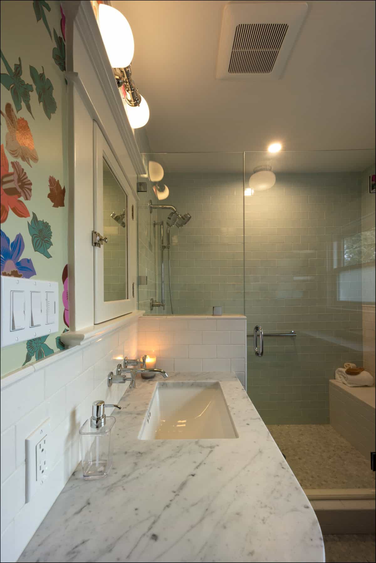 Modern bathroom with a marble countertop, sink, glass shower enclosure, wall mirror, and floral wallpaper on the left wall.