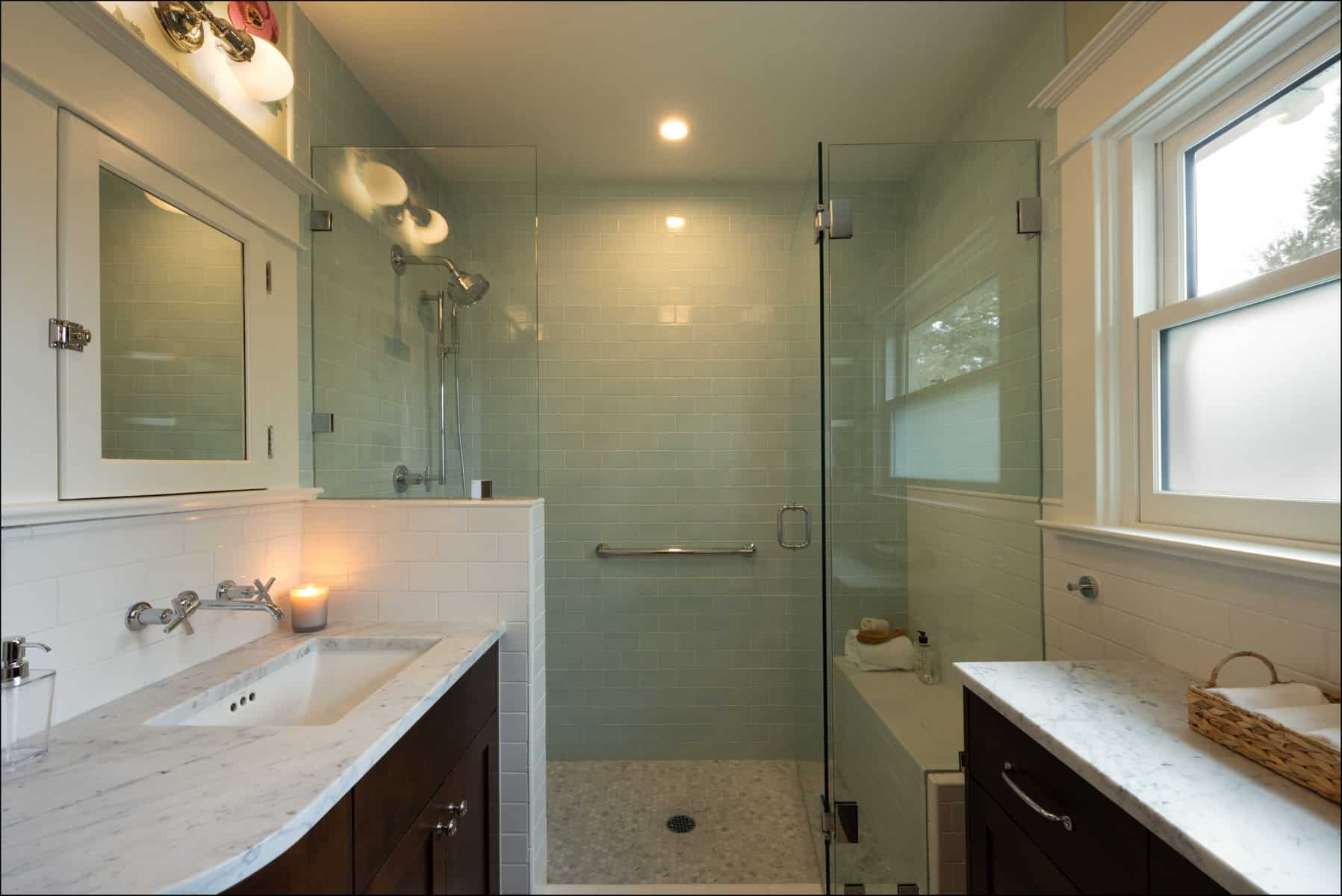 Modern bathroom with a glass-enclosed shower, white tile walls, a marble countertop sink, a lit candle, and a window letting in natural light.