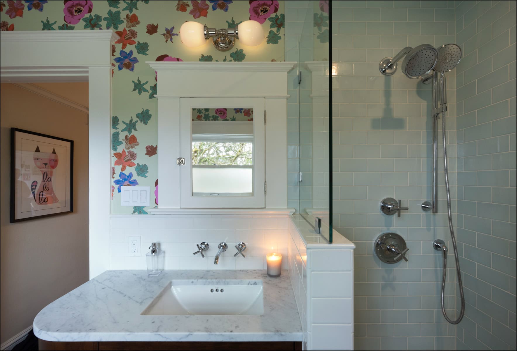 A bathroom with a marble sink, wall-mounted faucet, floral wallpaper, a lit candle, and a glass-enclosed shower with silver fixtures and a handheld showerhead.