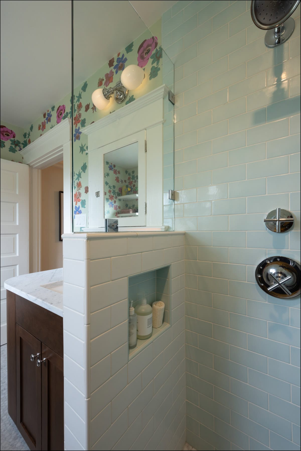 A bathroom with light blue subway tile in the shower, built-in shelf with toiletries, a vanity with marble countertop, and floral wallpaper on the upper wall.