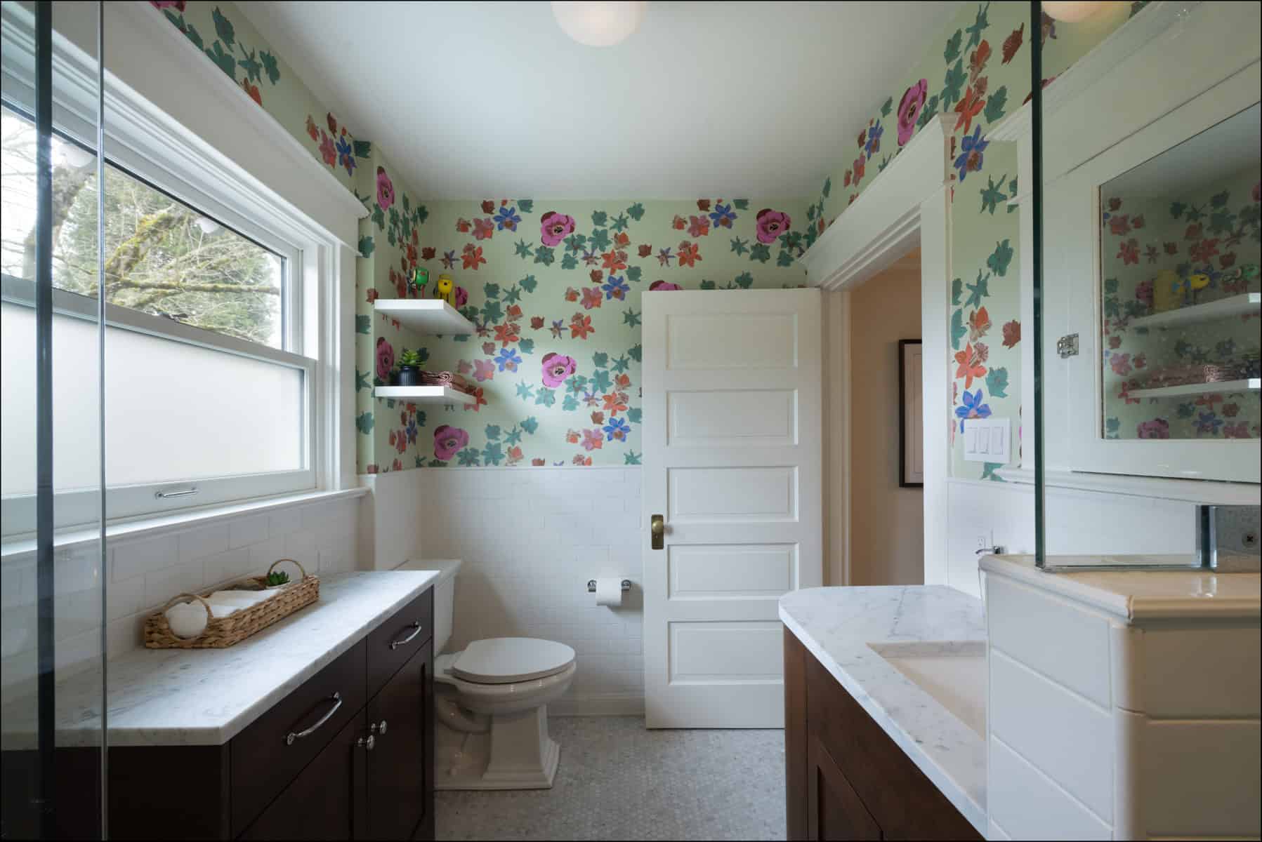 A bathroom with floral wallpaper, white tiled walls, a toilet, dark wood cabinets, marble countertops, and a window with frosted glass.