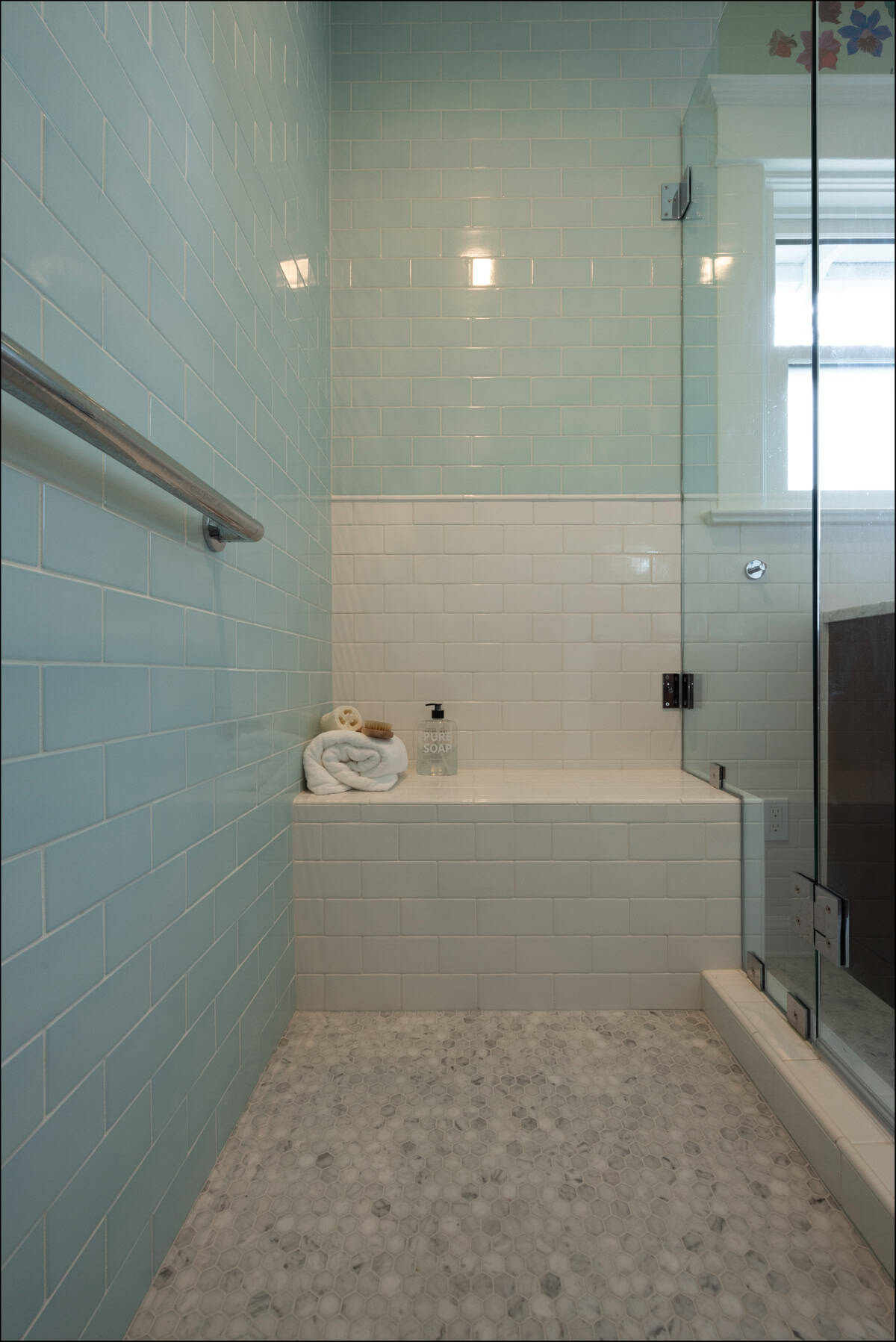 A tiled shower with light blue walls, white lower tiles, a built-in bench, a towel, soap dispenser, glass door, and a pebble-patterned floor.