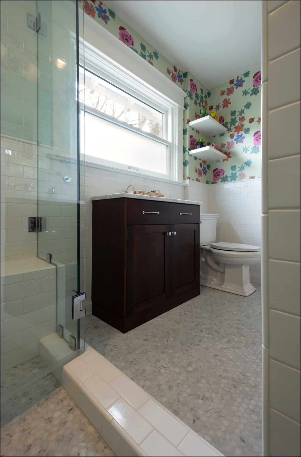 A bathroom with a glass shower, dark wood vanity, toilet, white tile, and colorful floral wallpaper above white wainscoting.