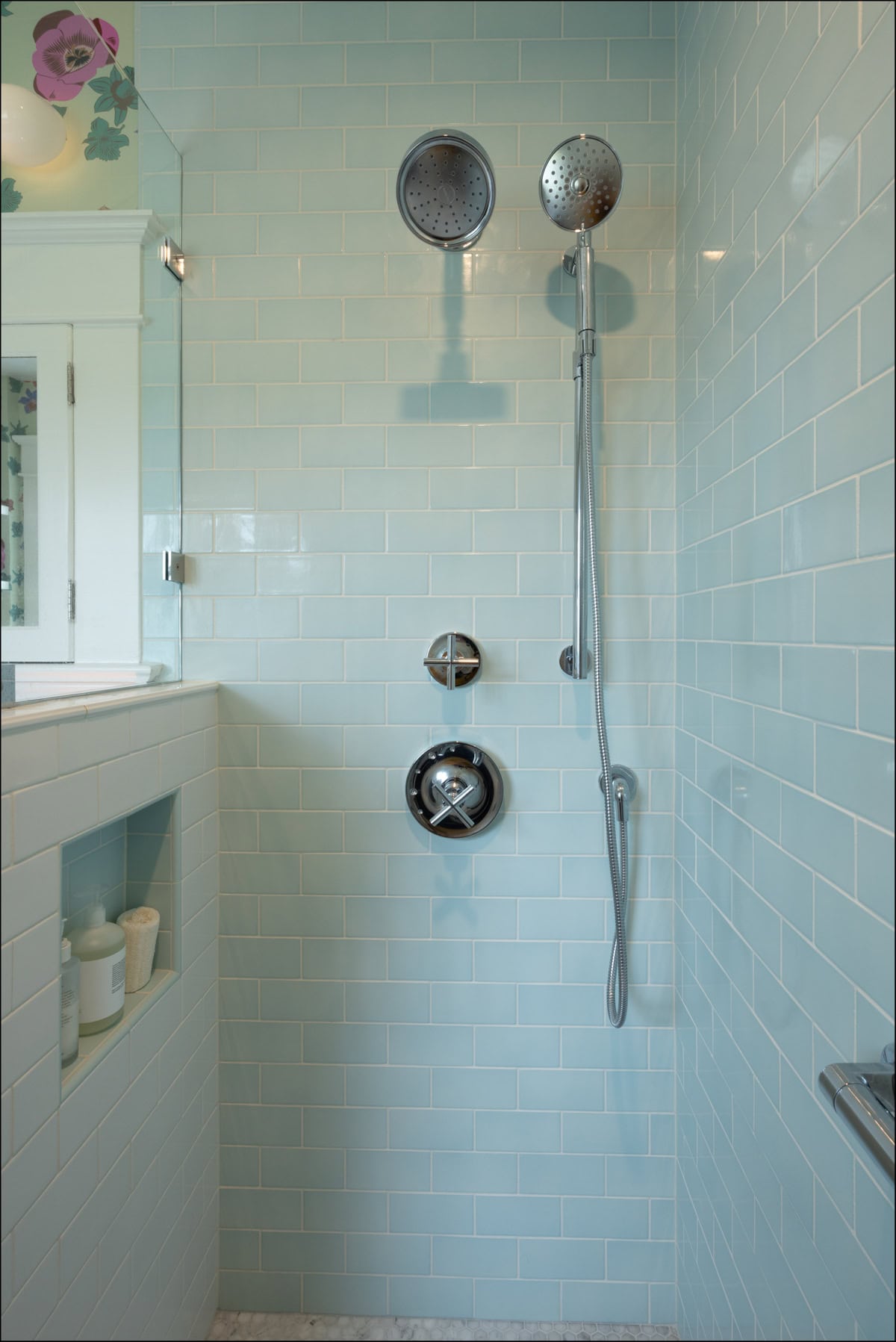 A modern shower with light blue subway tiles, two showerheads, a built-in shelf with bottles, and a glass door.