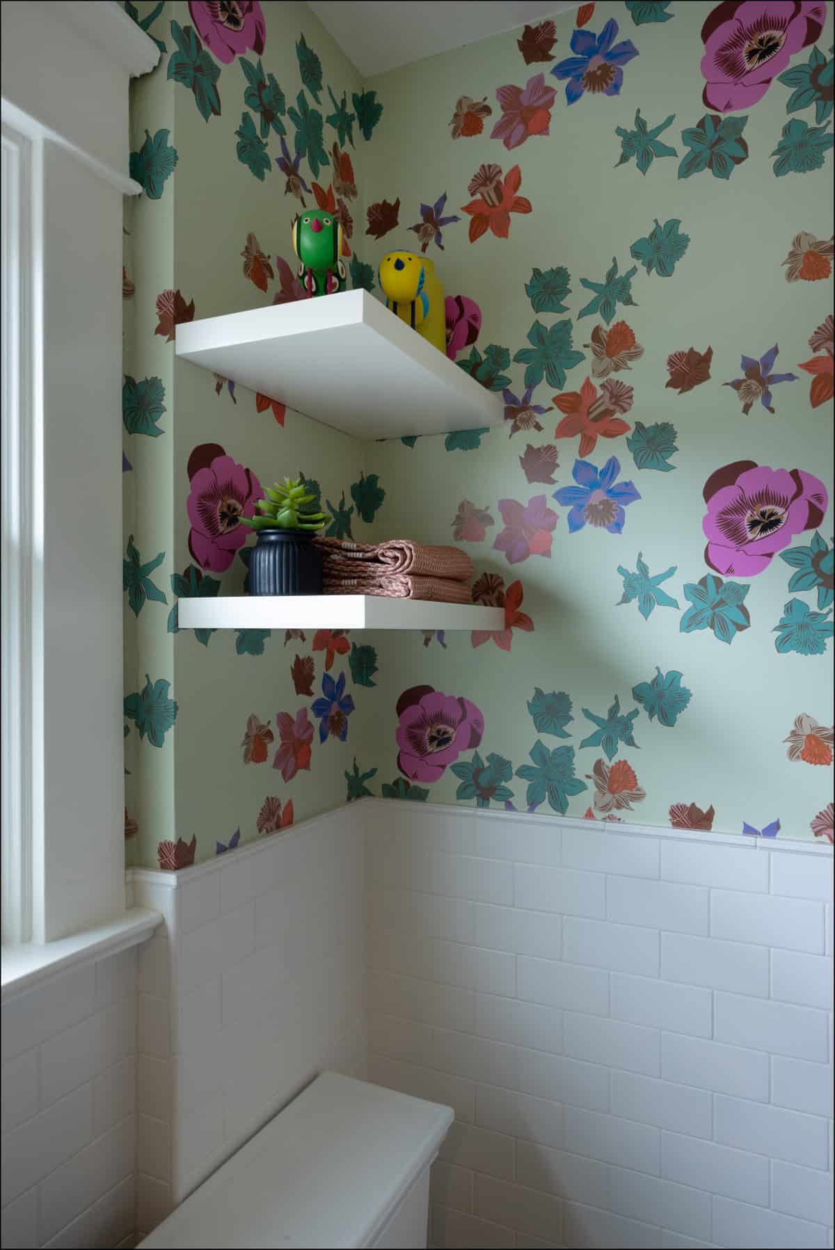 Two white shelves hold two bird figurines, a small plant, and folded towels against a floral wallpapered bathroom wall with white tile wainscoting.