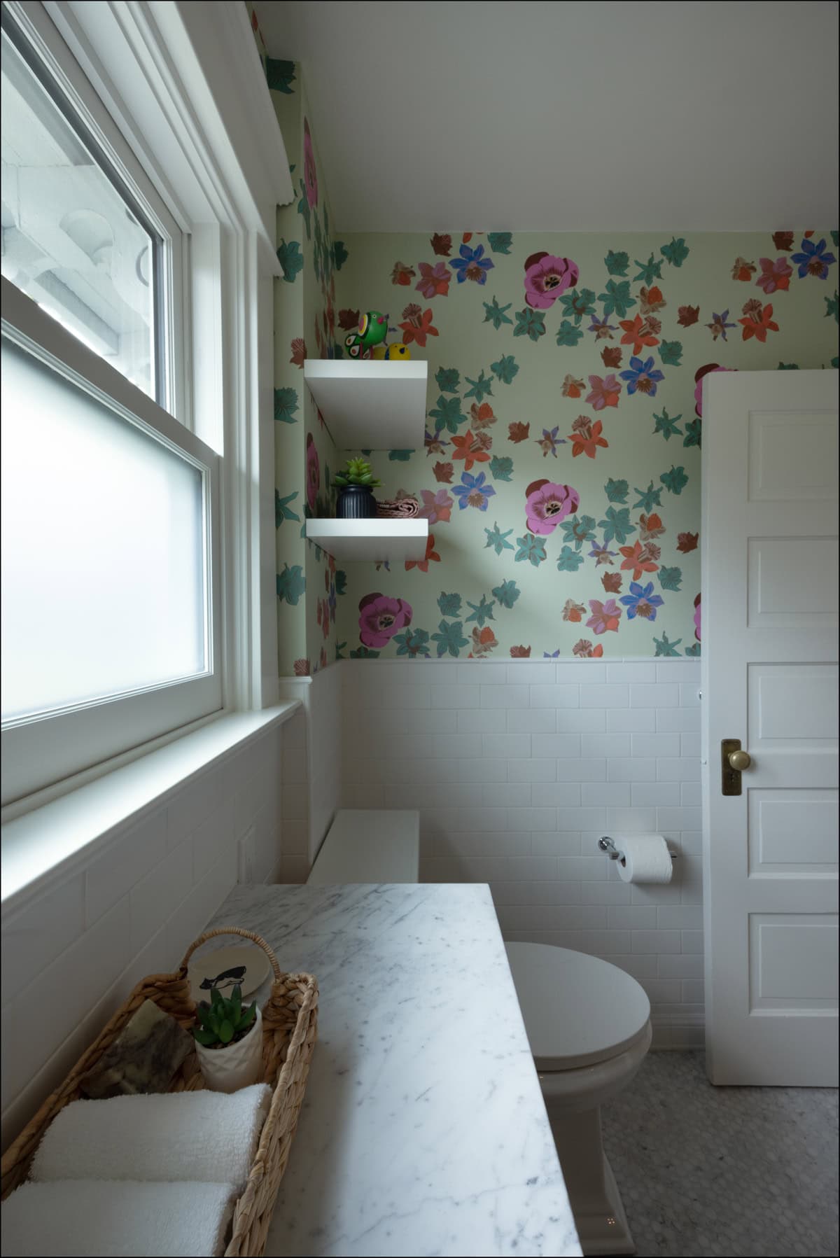 A small bathroom with floral wallpaper, marble countertop, wicker basket with towels, white tile walls, and shelves with plants above the toilet.