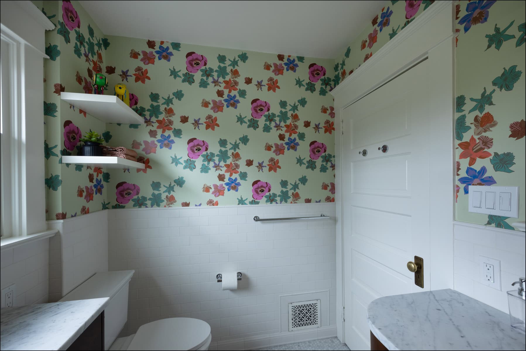 A bathroom with floral wallpaper, white tile walls, a toilet, floating shelves with decor, a marble countertop, and a white door.
