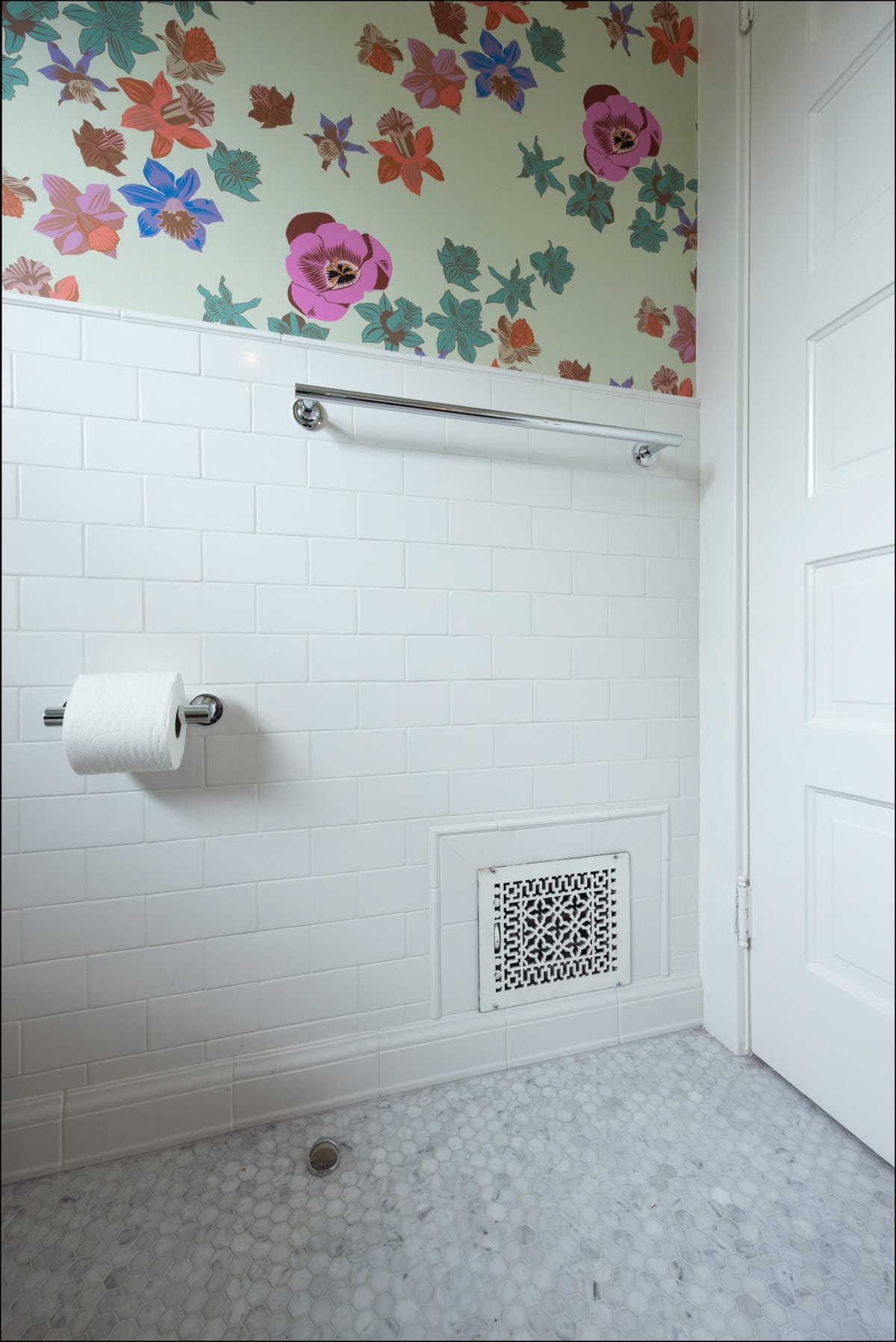 Bathroom wall with white subway tiles, floral wallpaper above, a towel bar, toilet paper holder with a roll, vent cover, and hexagonal tile floor.