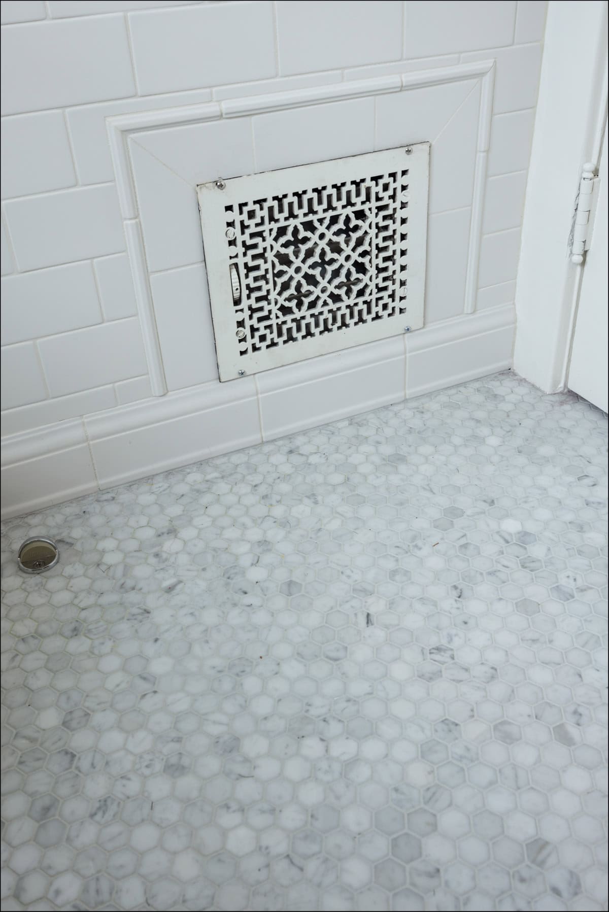 White tiled bathroom wall with a decorative vent cover, hexagonal marble floor tiles, and a round floor drain visible in the bottom left corner.