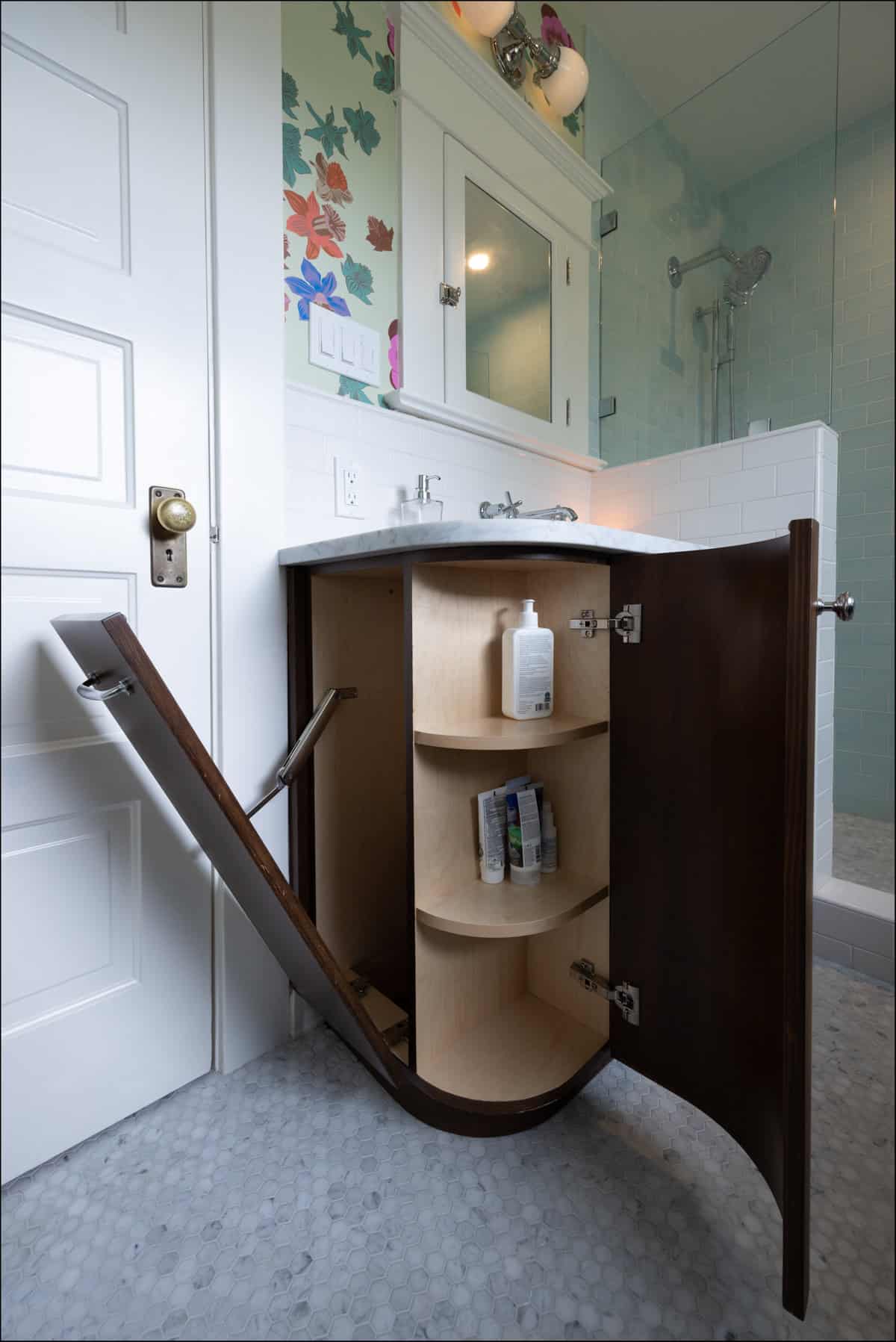 Bathroom with a curved wooden vanity cabinet door open, revealing shelves with toiletries inside; sink and mirror above, shower visible in the background.