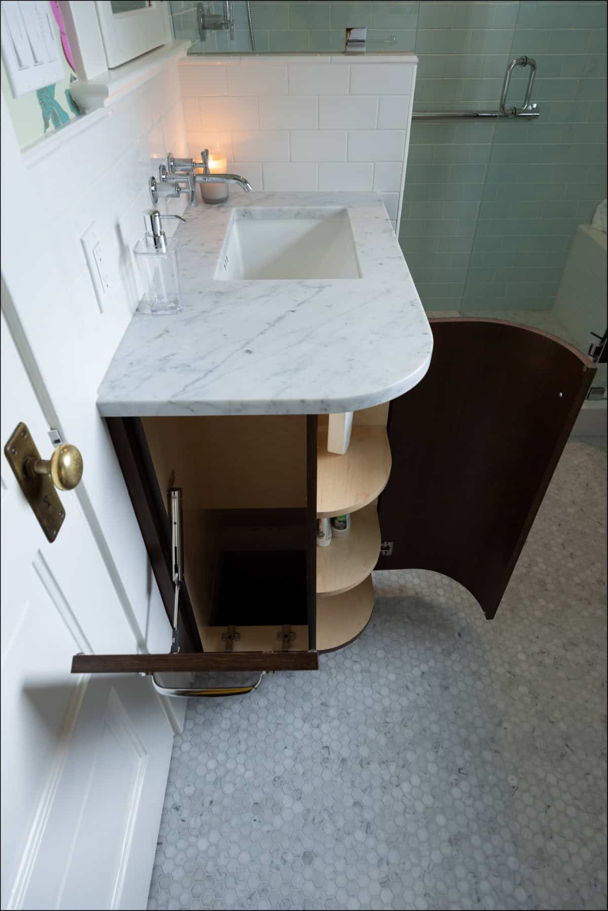 A bathroom vanity with marble countertop, open cabinet and drawer revealing empty shelves and plumbing, next to a sink and soap dispenser.