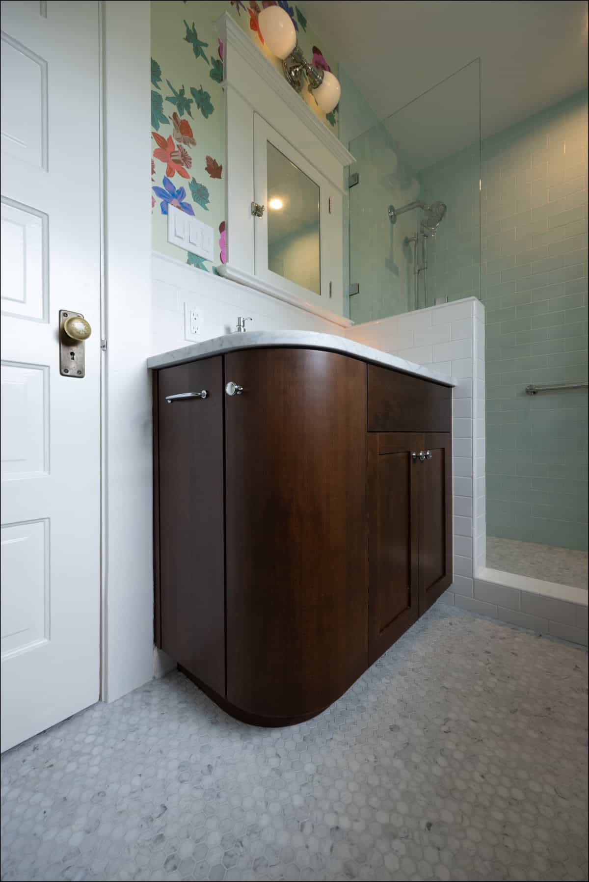 A bathroom with a dark wood vanity, white countertop, floral wallpaper, and a glass-enclosed shower with white tile.