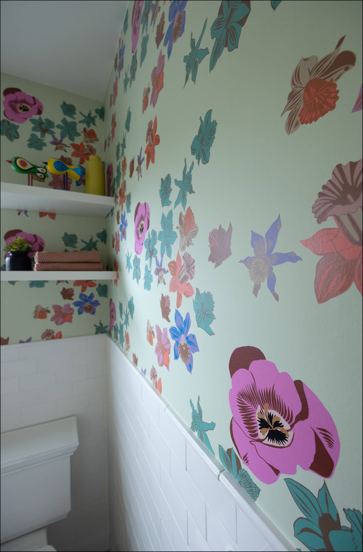Bathroom wall with colorful floral wallpaper, white subway tile, and white shelves holding books, plant, and decorative items.