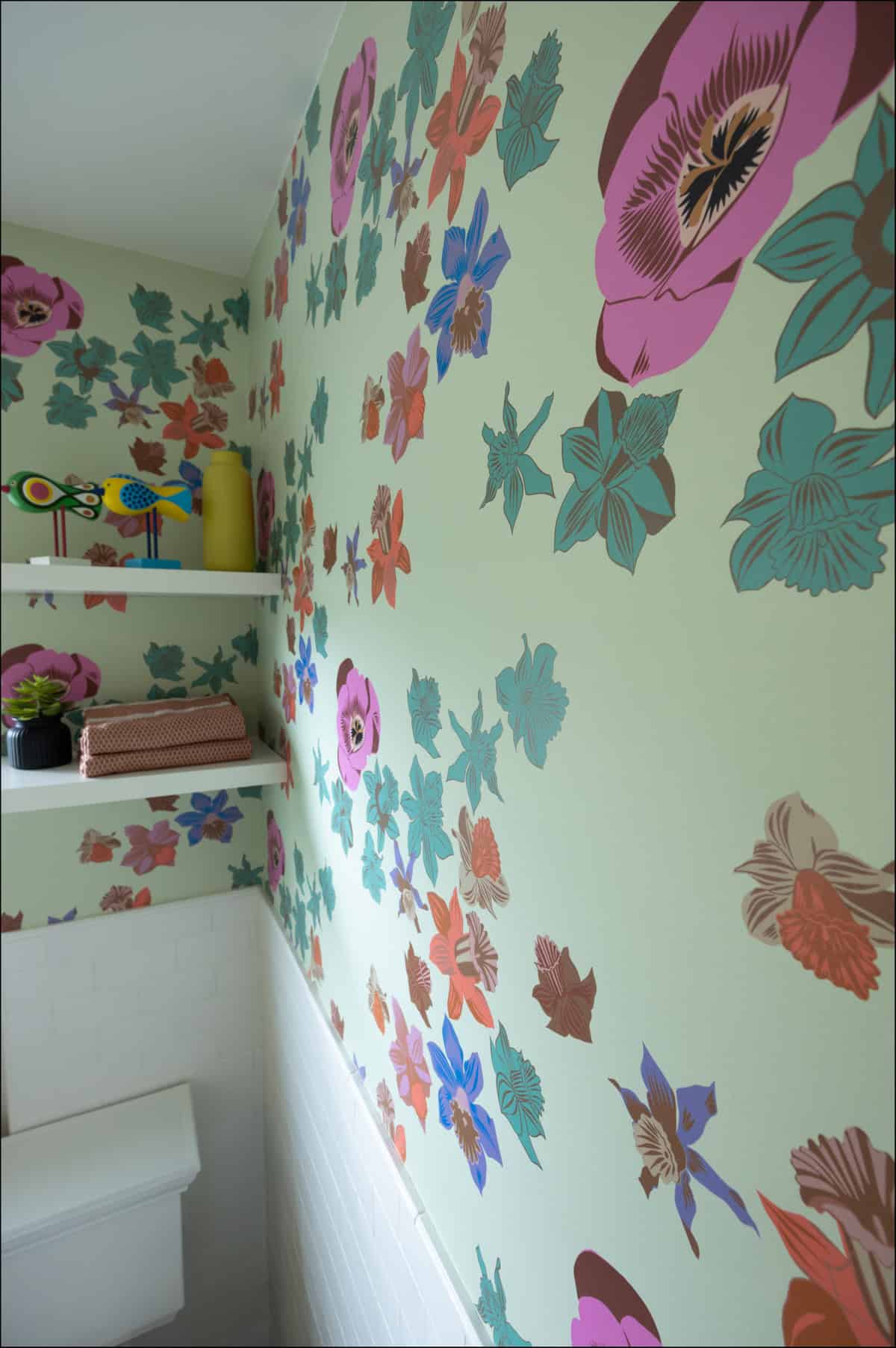 A bathroom wall decorated with colorful floral wallpaper, two white shelves holding folded towels, a plant, a vase, and decorative bird figures.