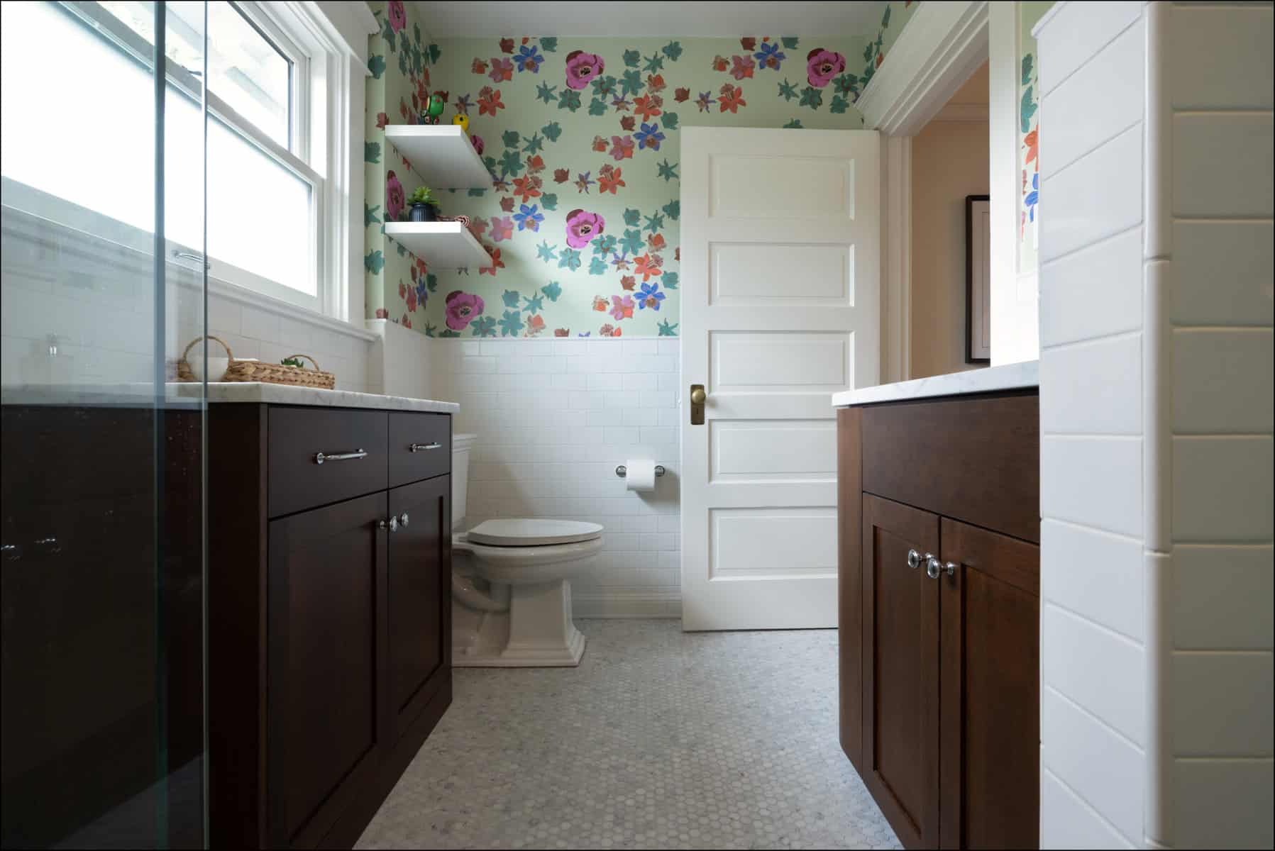 A bathroom with floral wallpaper, dark wood cabinets, white countertops, a toilet, two floating shelves, and a window letting in natural light.