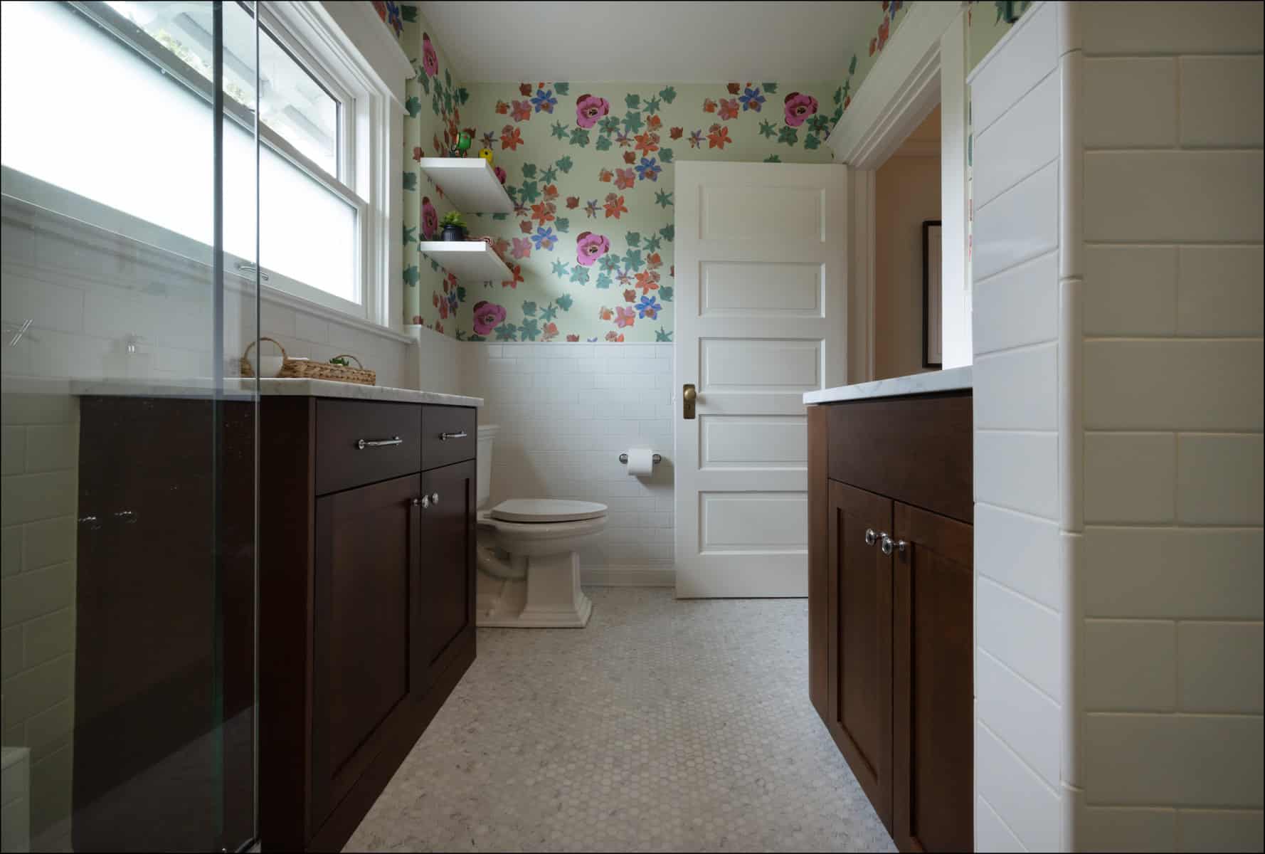A bathroom with floral wallpaper, dark wood cabinets, a toilet, white tile walls, and a window above the sink.