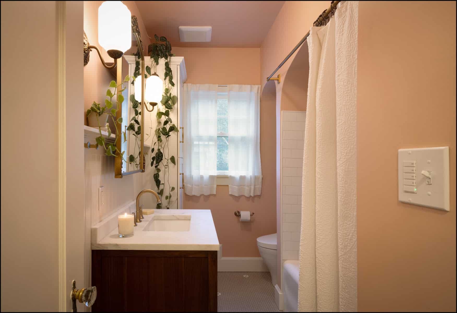 A small bathroom with peach walls, a white sink vanity, a mirror, hanging plants, a lit candle, a curtained window, and a shower with a white curtain.