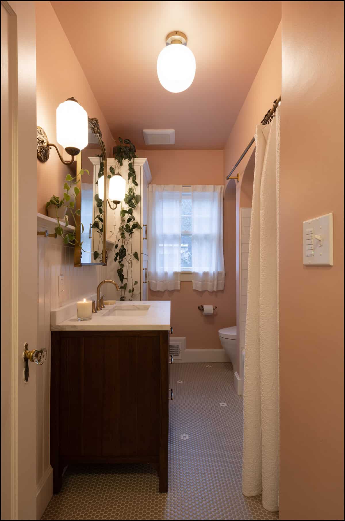 A bathroom with pink walls, a wooden vanity, a lit candle, hanging plants, a window with white curtains, a toilet, and a shower with a white curtain.