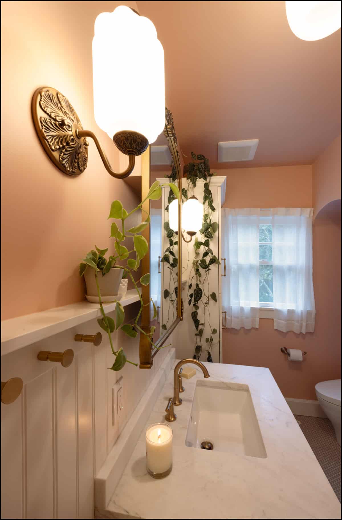 A bathroom with pink walls, a white sink, brass fixtures, a lit candle, a wall-mounted plant, a mirror, and natural light coming through a window with sheer curtains.