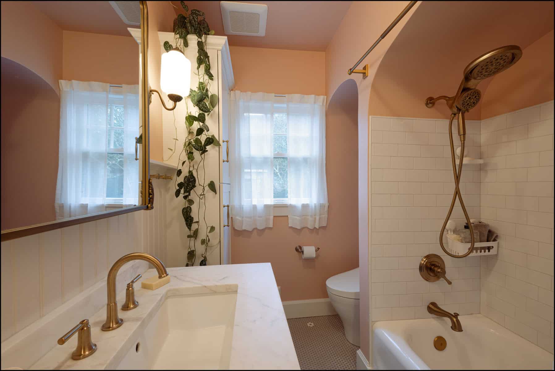 A bathroom with pink walls, white tile, brass fixtures, a marble sink, a bathtub with a shower, a toilet, a hanging plant, and sheer curtains on the window.