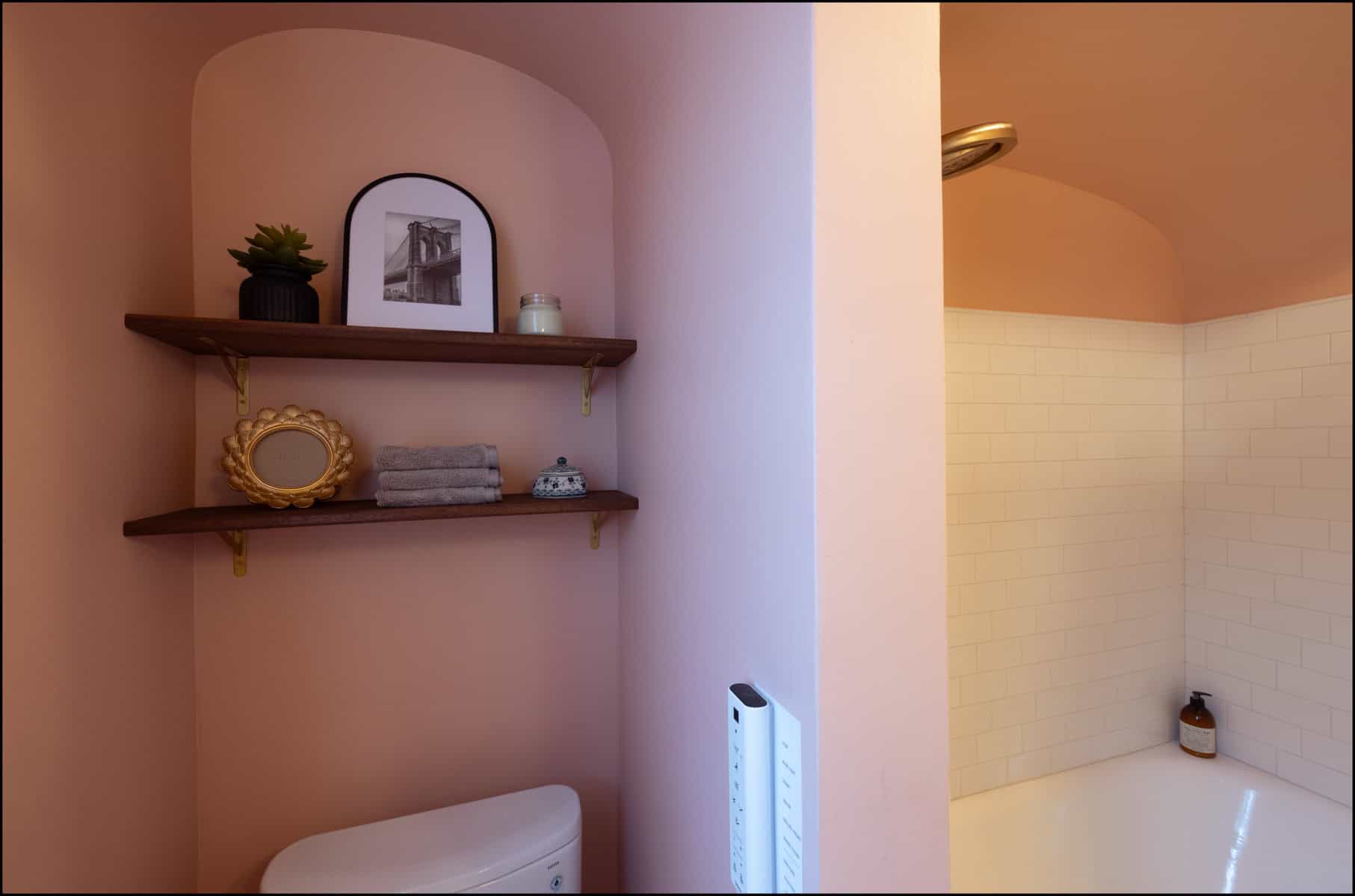 Bathroom with peach-colored walls, wooden shelves holding decor and towels above a toilet, and a shower with white tile partially visible.