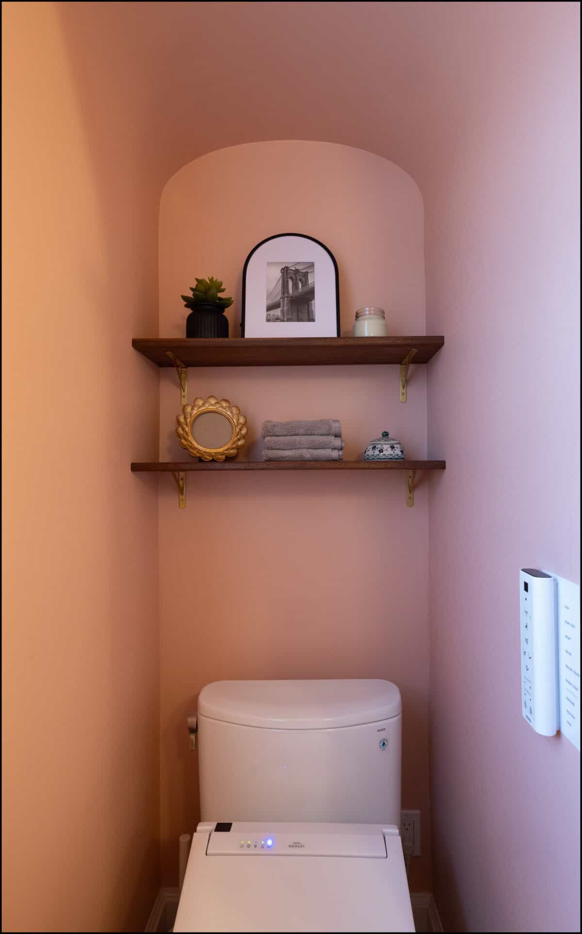 A modern toilet sits below two wooden shelves holding a framed photo, plant, candle, mirror, folded towels, and a small container against a peach-colored wall.