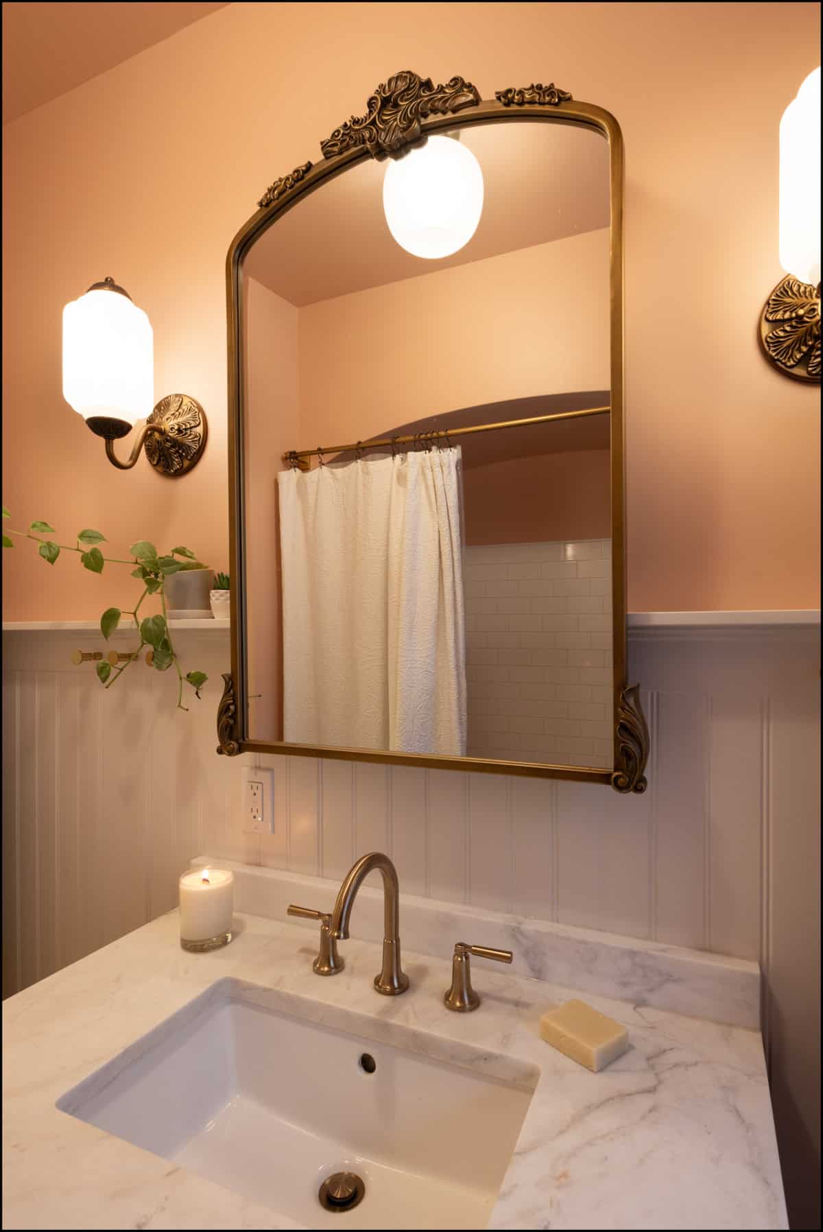 A bathroom vanity with a marble countertop, gold faucet, soap, lit candle, plant, and a large ornate mirror reflecting a shower curtain.