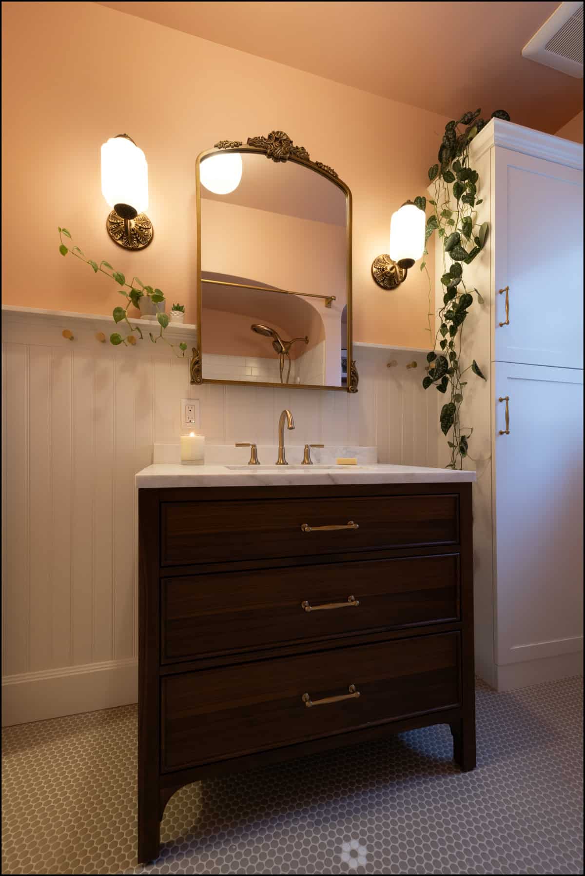 A bathroom vanity with a dark wood cabinet, brass fixtures, a rectangular mirror, wall sconces, and trailing plants against peach walls and white wainscoting.